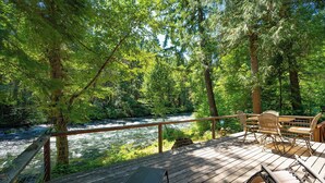 A wooden deck with outdoor furniture overlooking a river, surrounded by dense forest. - McKenzie Bridge River's Edge Retreat, a cozy cabin on over two acres (McKenzie Bridge)