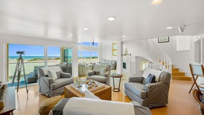 A spacious living area with multiple seating options, a staircase, and large windows offering a view of the beach. - Beachfront Villa w/ Direct Beach Access & Elevator!  (Oxnard)