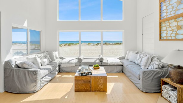 A spacious living room with a large window overlooking a beach, a grey sectional sofa, and a wooden coffee table. - Beachfront Villa w/ Direct Beach Access & Elevator!  (Oxnard)