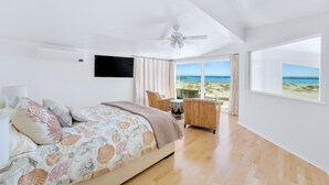 A bedroom with a bed, a ceiling fan, a TV mounted on the wall, a wicker chair, and a view of the beach through large windows. - Beachfront Villa w/ Direct Beach Access & Elevator!  (Oxnard)