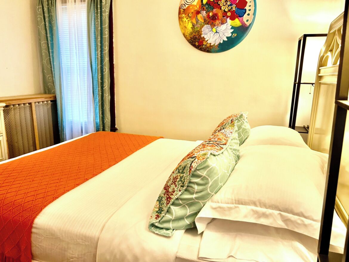 A neatly made bed with a floral pillow and a red blanket, a radiator, and a wall with a floral artwork.
