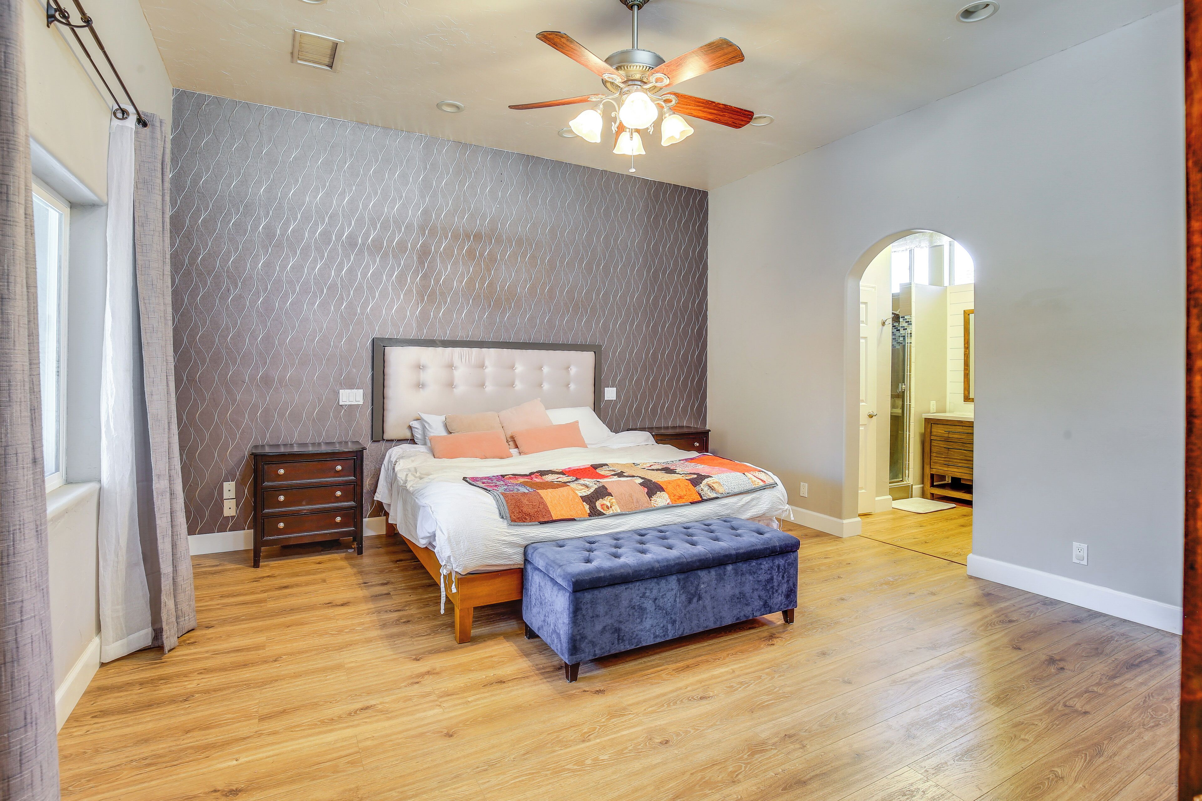 A bedroom with a wooden floor, a bed with a white headboard, a blue bench, and a ceiling fan.