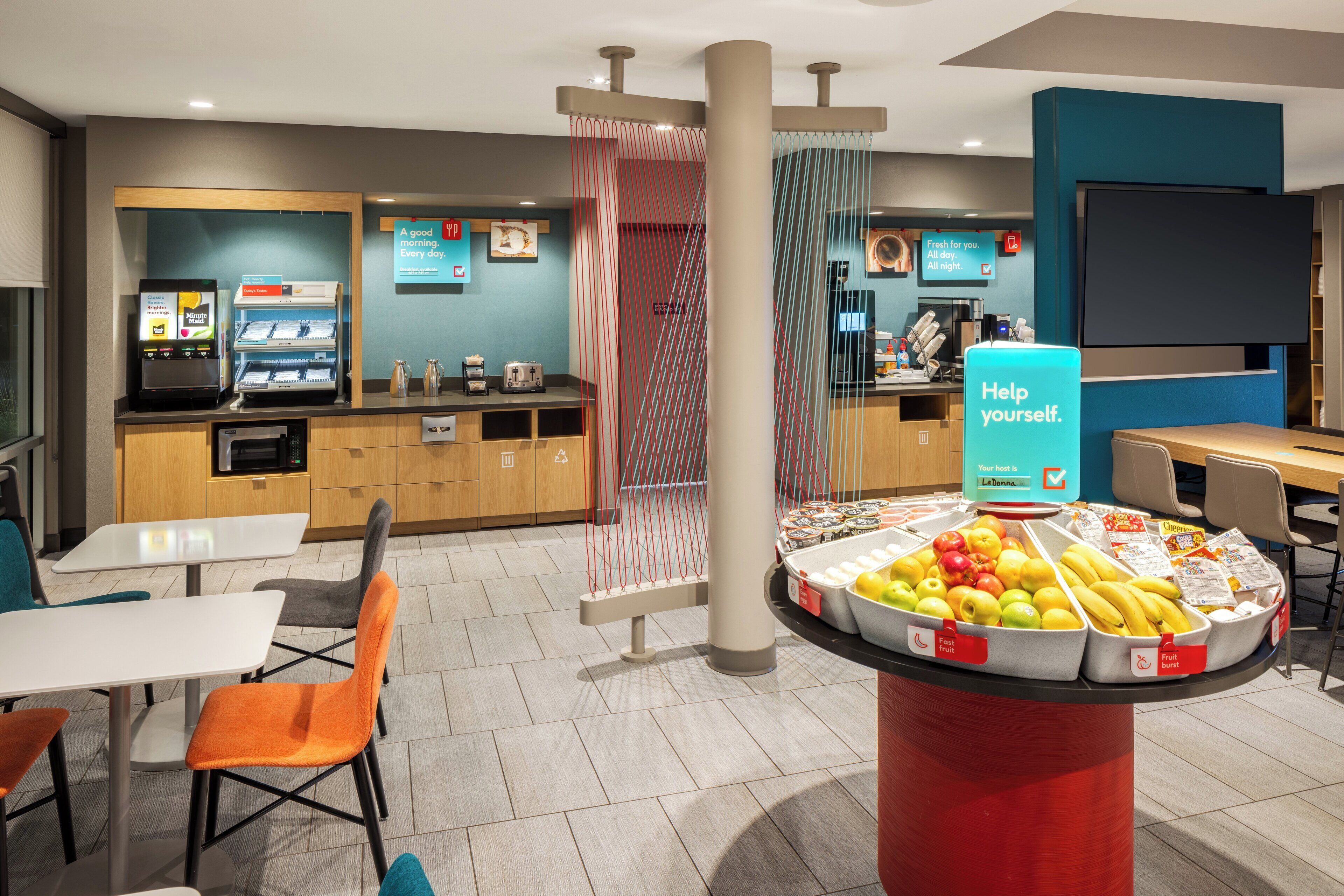 Un moderno vestíbulo de hotel con una estación autoservicio de frutas y bebidas, áreas de estar y una máquina de café.