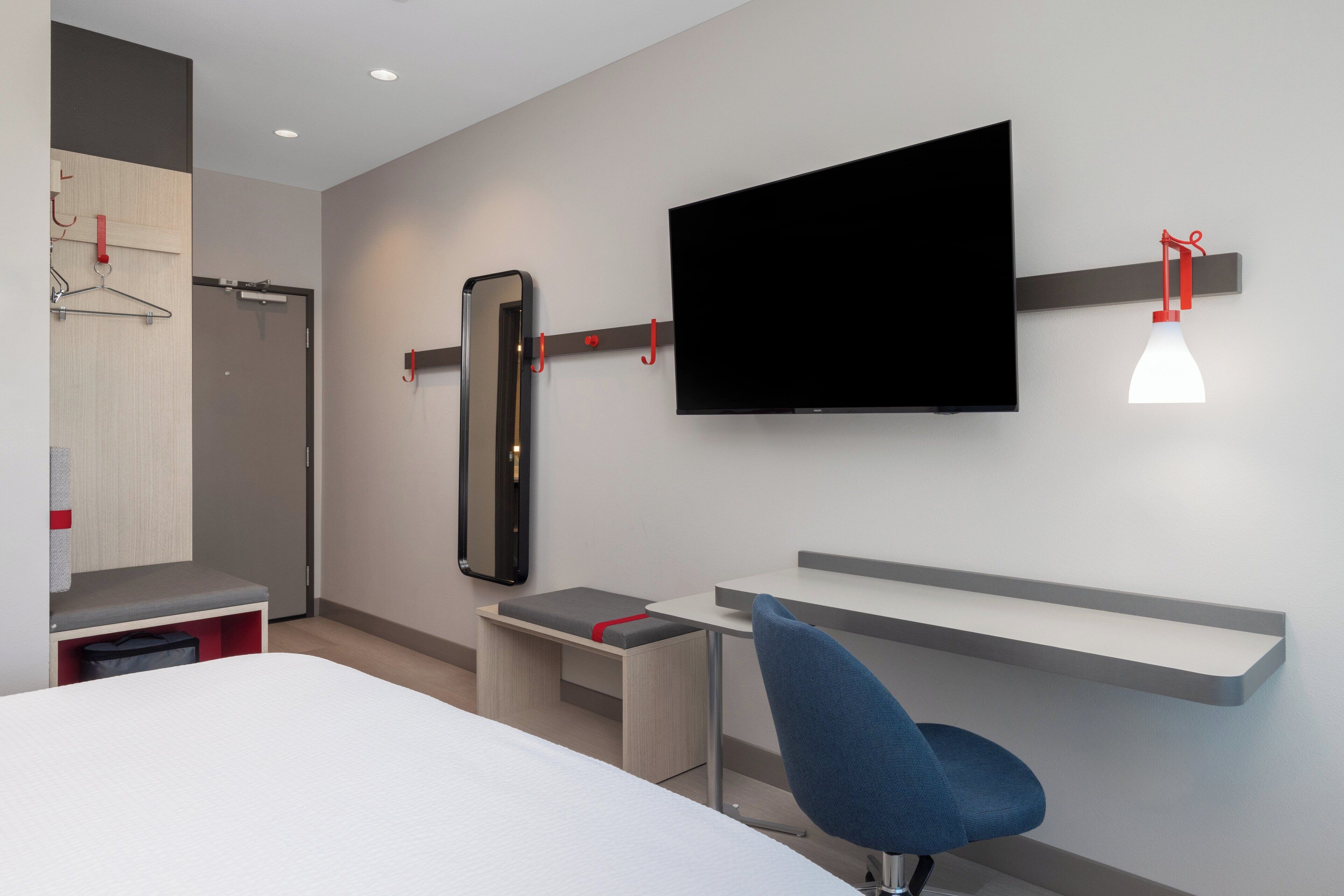 A modern hotel room with a bed, a desk, a chair, a TV mounted on the wall, and a mirror.