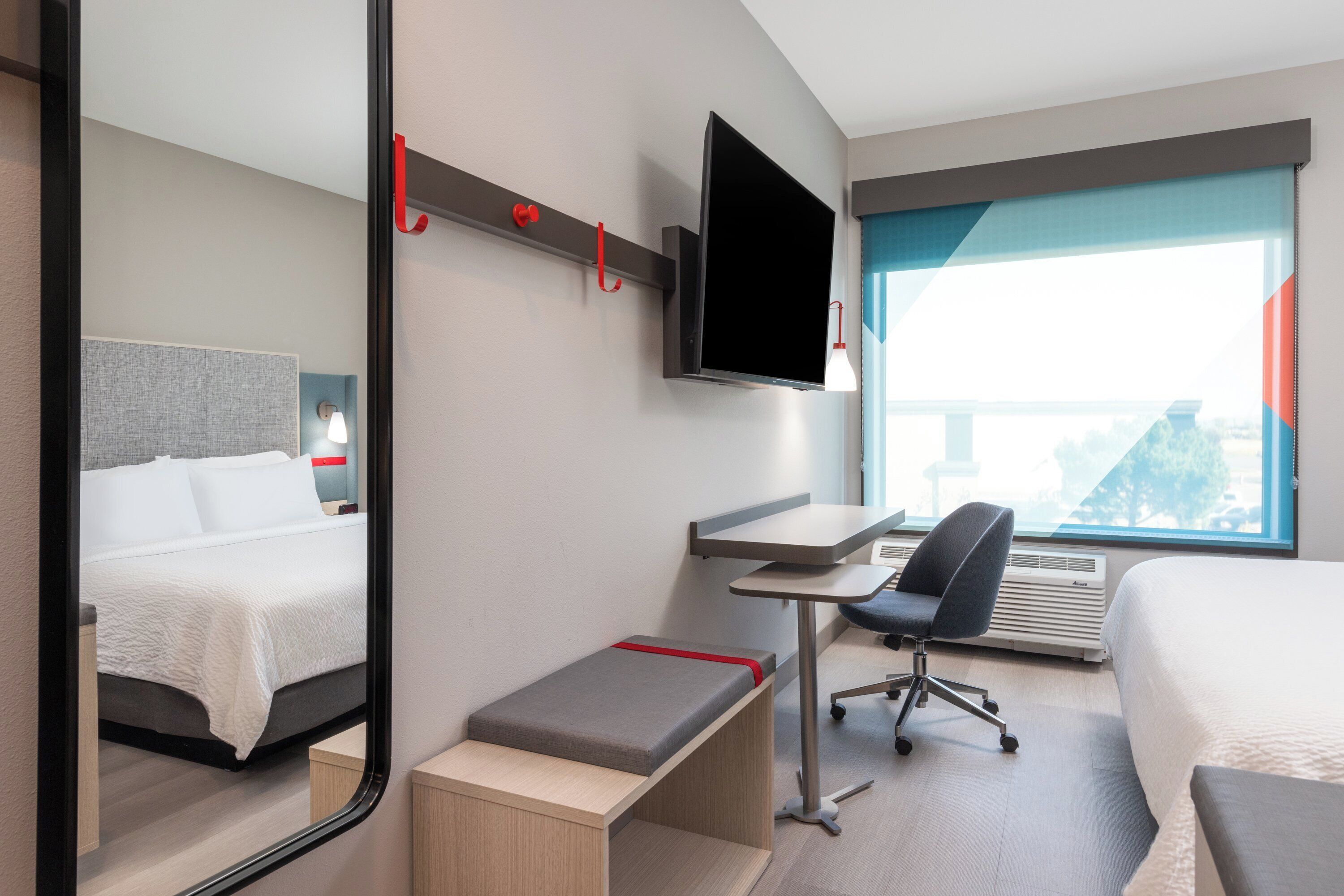 A hotel room with a bed, a desk with a chair, a TV mounted on the wall, and a large window offering a view.