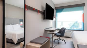 A hotel room with a bed, a desk with a chair, a TV mounted on the wall, and a large window offering a view.