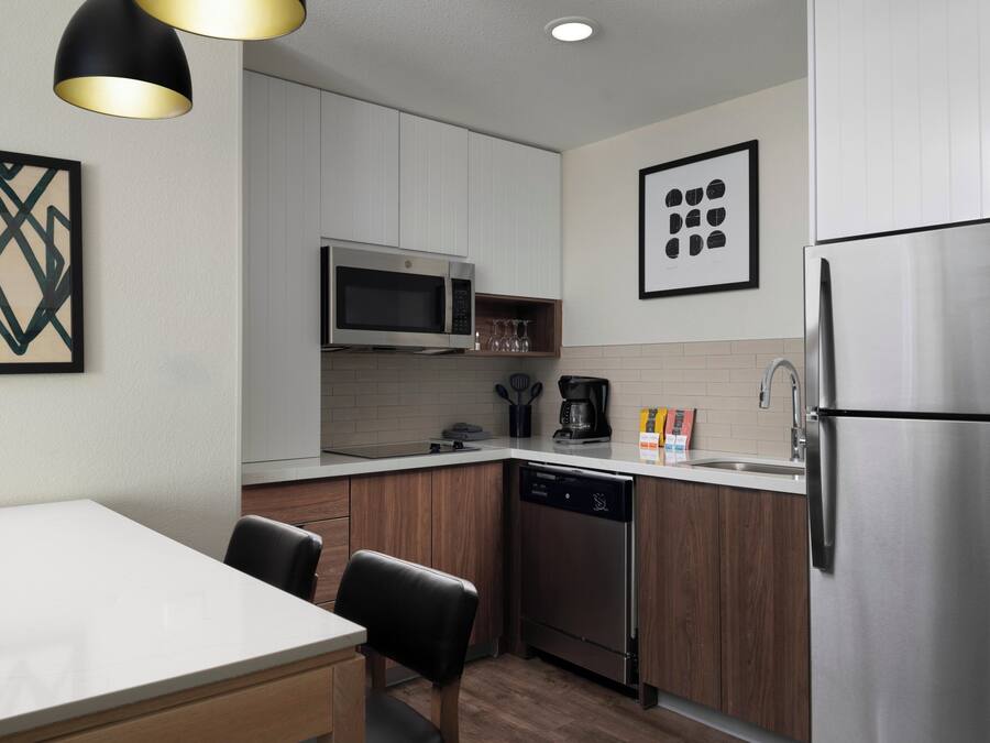 A compact kitchen with white cabinets, a stainless steel refrigerator, a microwave, a coffee maker, and a dishwasher. There are two chairs and a table.