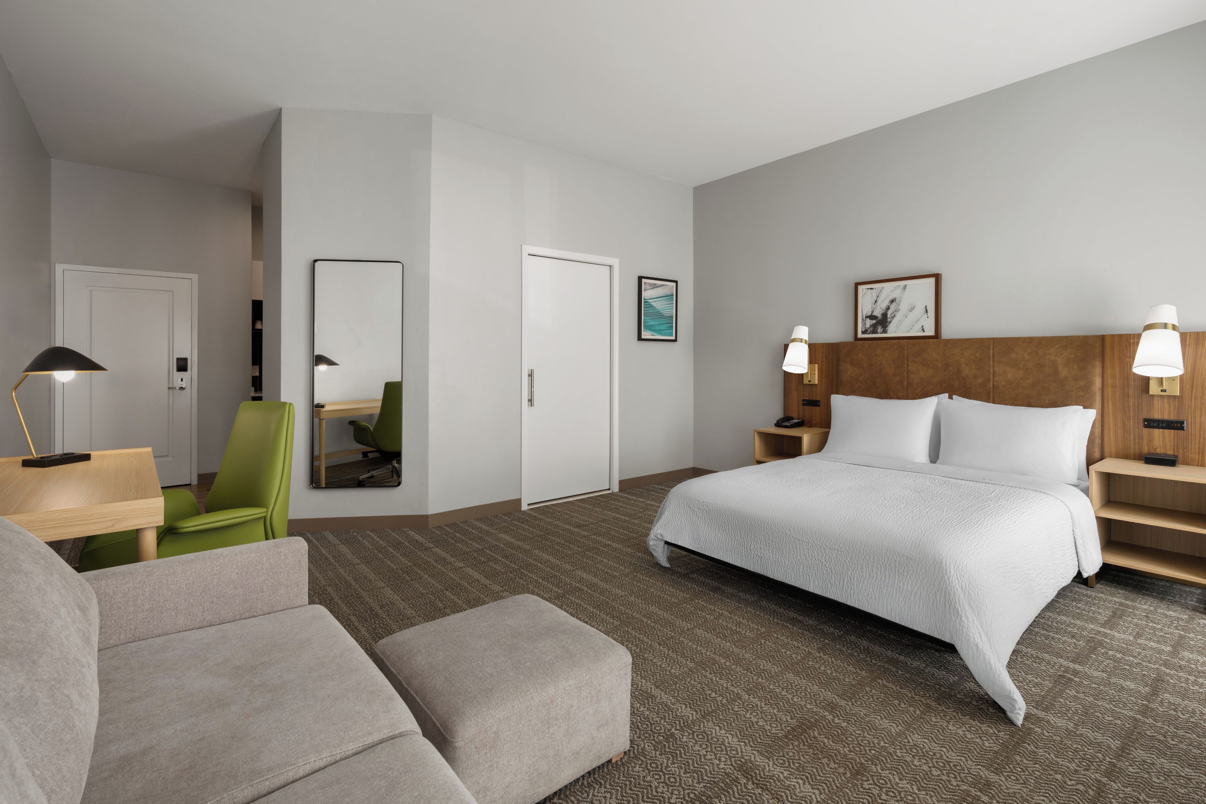 A hotel room with a bed, a desk, a chair, a sofa, a mirror, and a nightstand with a lamp.