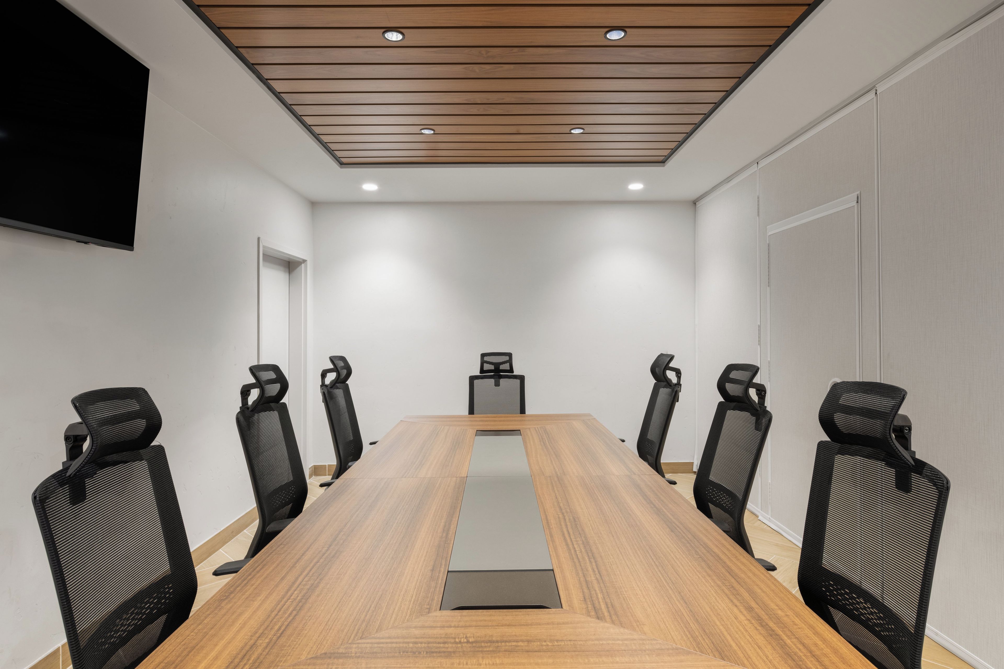 Une salle de conférence avec une longue table en bois, des chaises noires à dossier en résille, des murs blancs et un éclairage encastré au plafond.