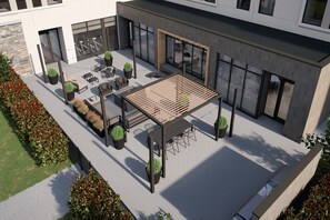 Un espace extérieur aménagé en patio, avec des sièges et des jardinières, entouré d’un bâtiment doté de grandes fenêtres et d’un mur en pierre.