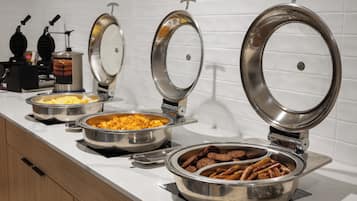 A buffet setup with various food items in metal containers, a waffle maker, and a coffee machine.