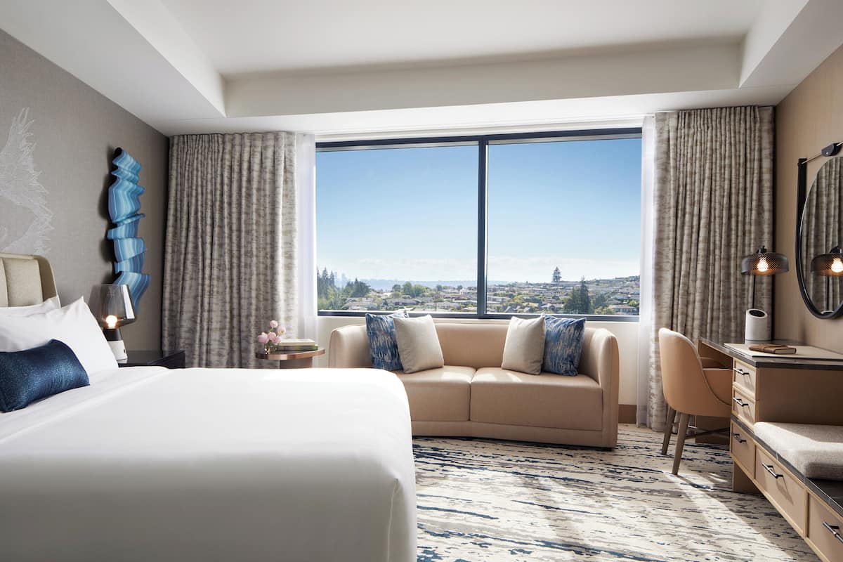 A hotel room with a large bed, a sofa, a desk, and a view of a cityscape through a large window.