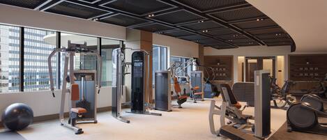 A modern gym with various exercise equipment, large windows, and a high ceiling.