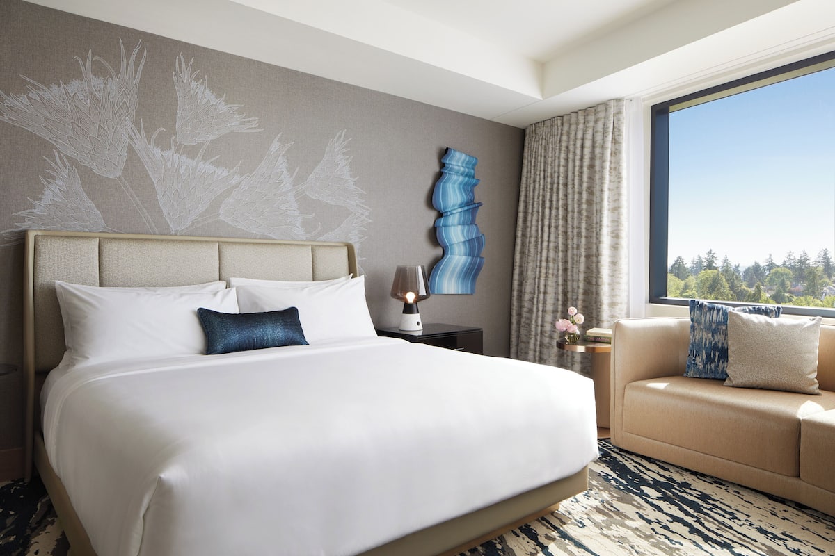 A hotel room with a large bed, a sofa, a nightstand with a lamp, a decorative wall piece, and a window with a view of trees.