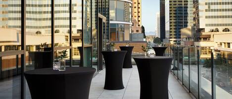 Terraço no telhado com mesas redondas pretas, grandes vasos de plantas e vista para prédios da cidade.