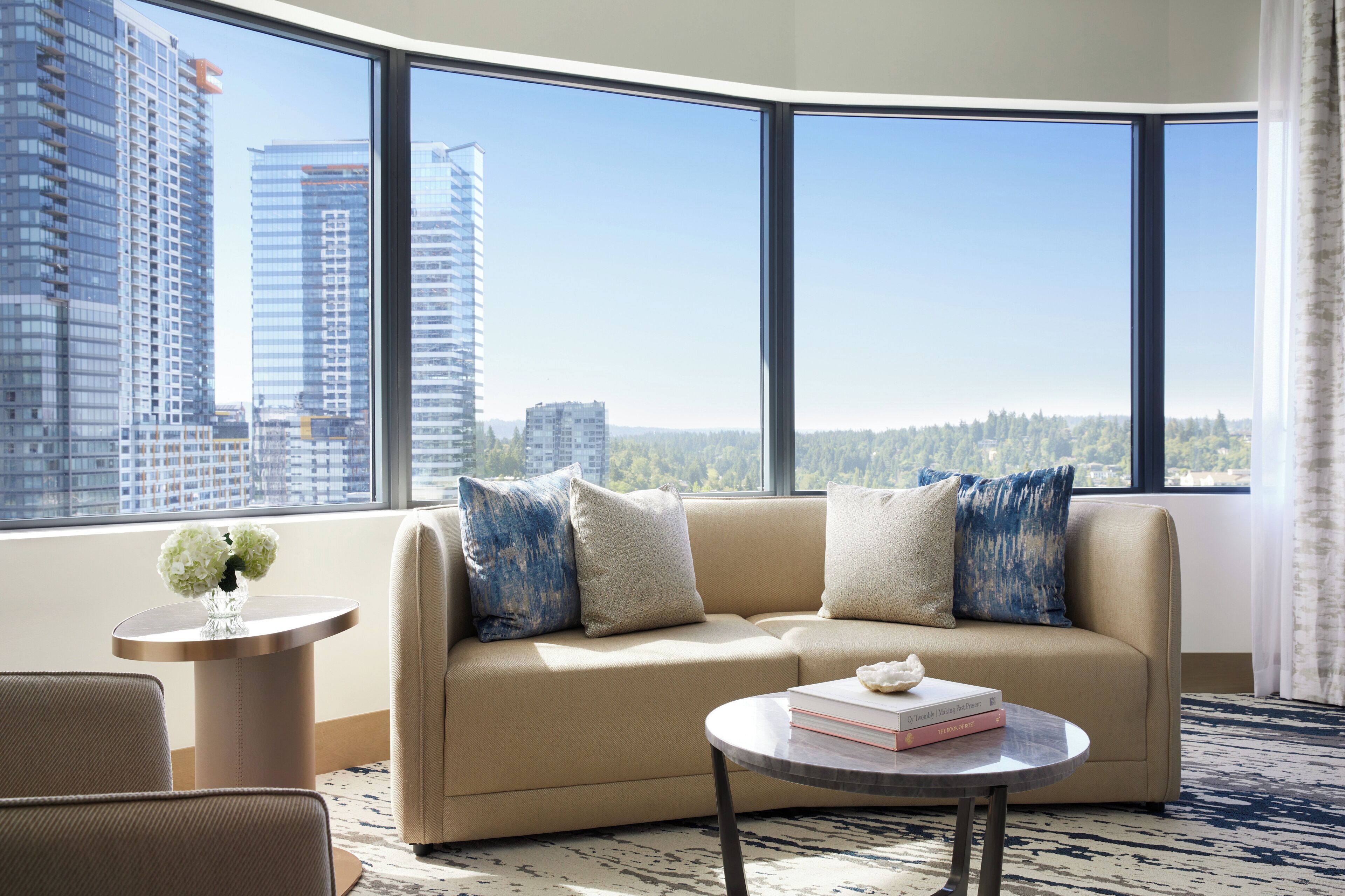 A modern living room with a beige sofa, a round coffee table, and a view of tall buildings through large windows.