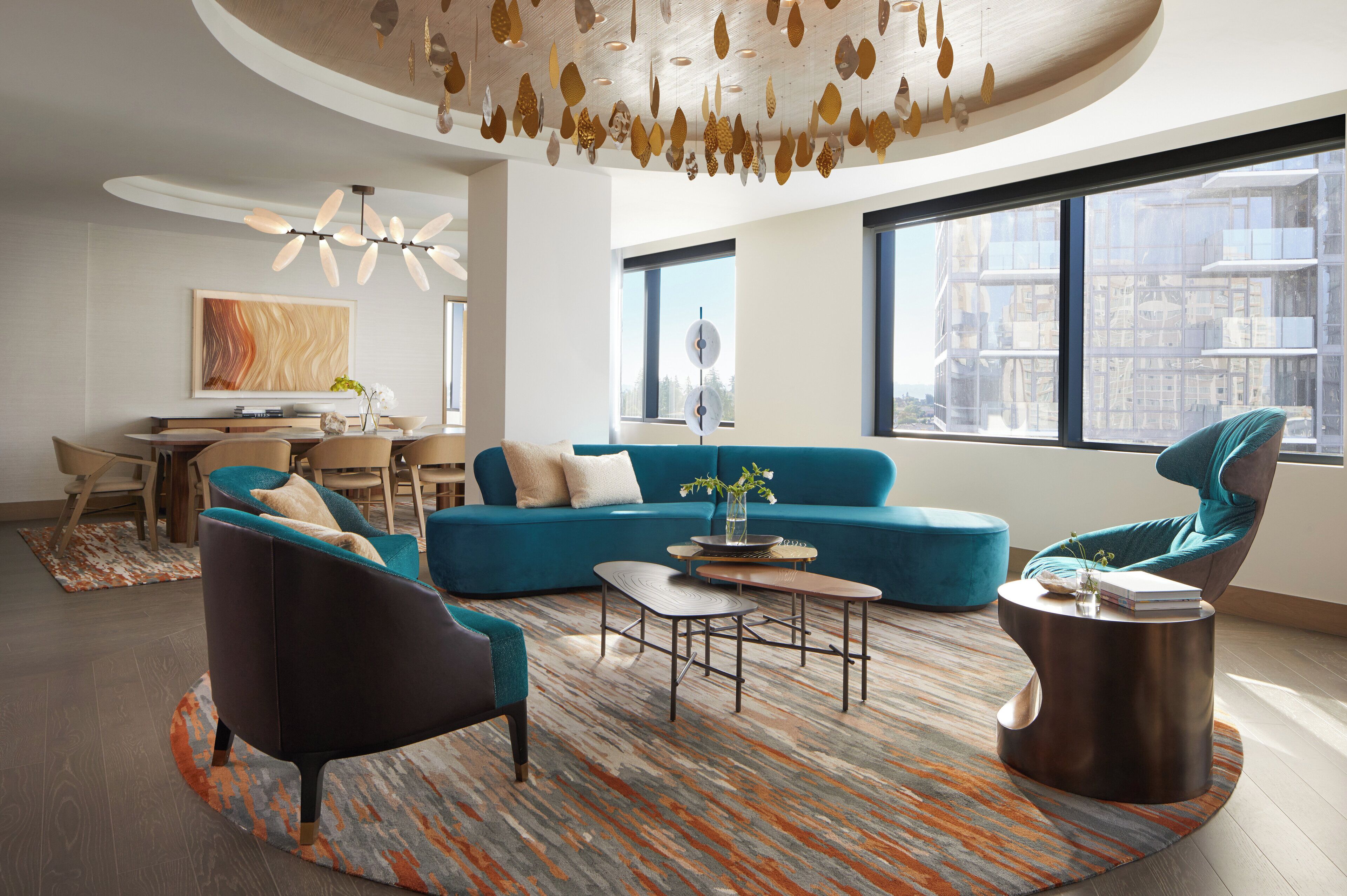 Un salon moderne avec un canapé bleu, un fauteuil foncé et une table basse ronde. On y trouve également un coin repas avec une table et des chaises en bois, ainsi qu’une grande fenêtre donnant sur la ville.