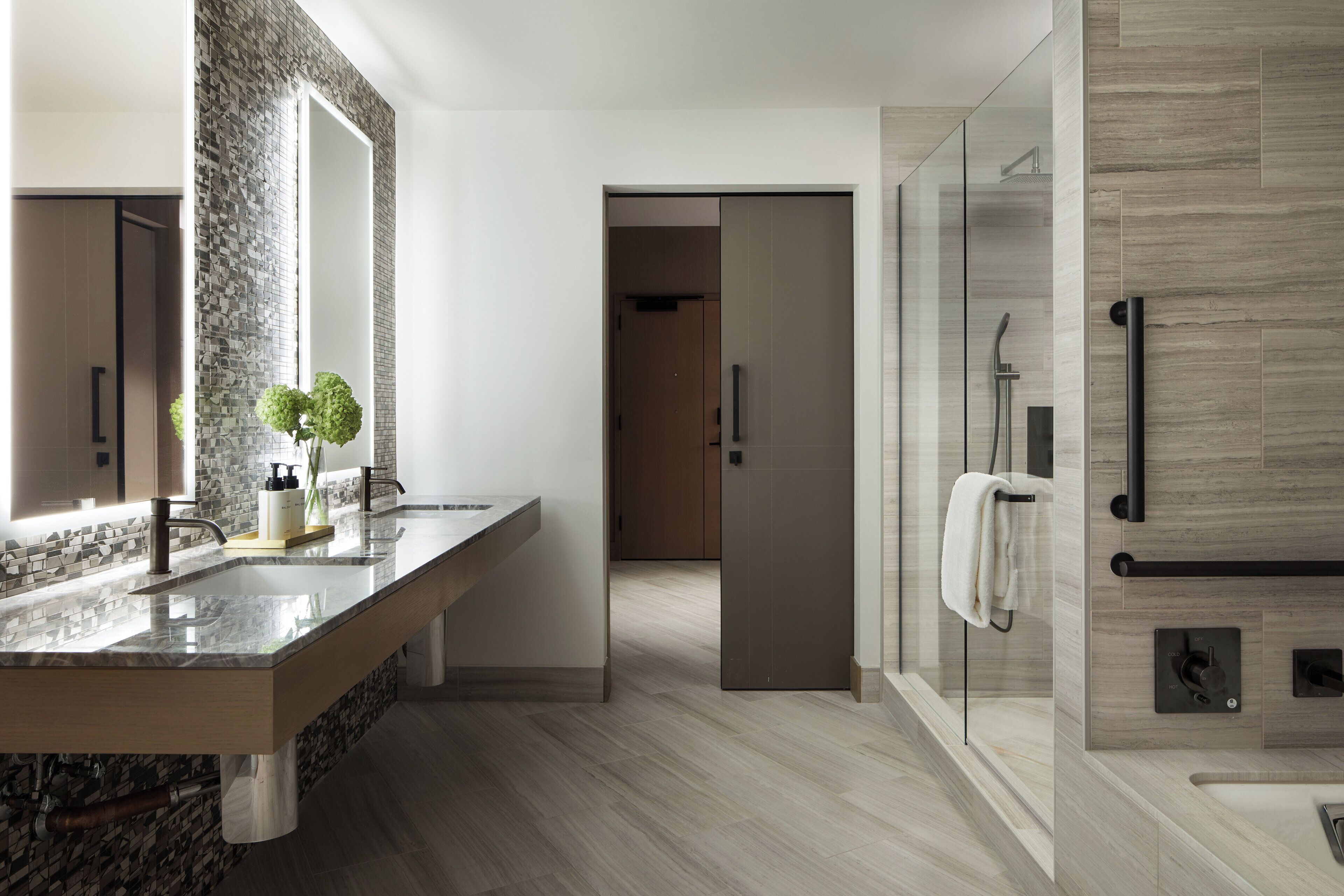 Un baño moderno con un espejo grande, lavamanos dobles, cabina de ducha con mampara de vidrio y tocador de madera.