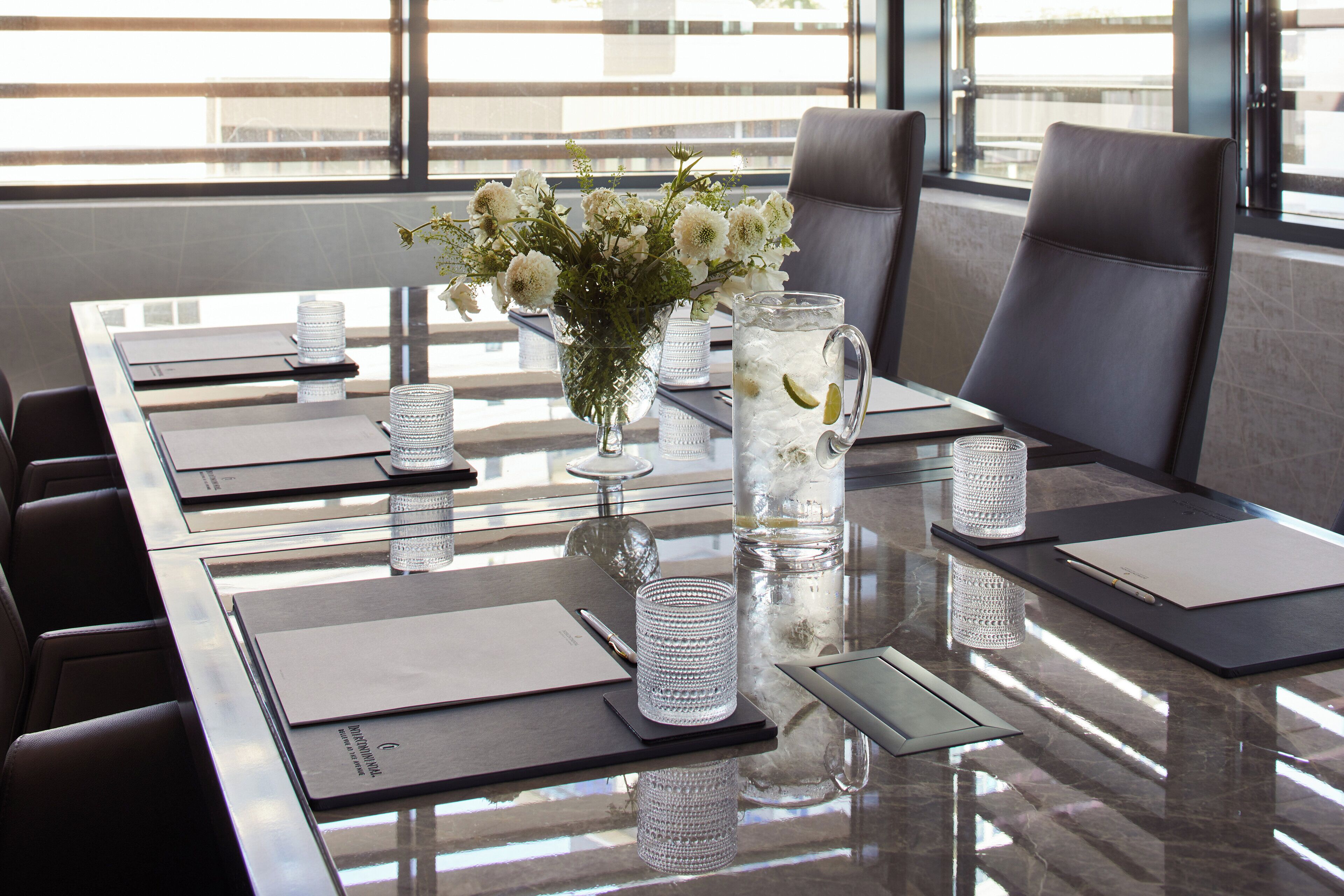A conference table with a vase of flowers, a pitcher of water with lime, and a glass of water.