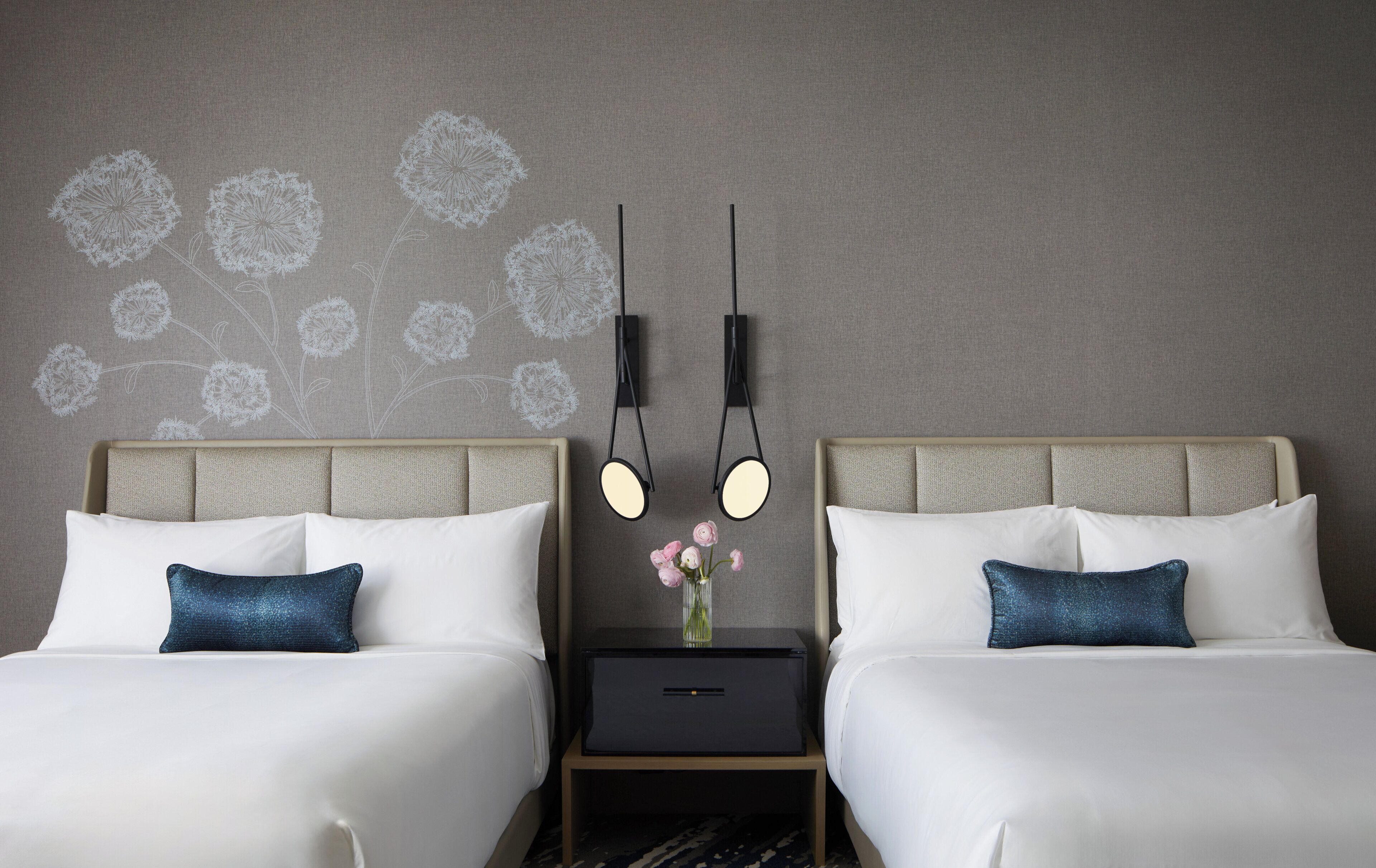 Habitación de hotel con dos camas, una mesita de noche con un jarrón de flores y lámparas fijadas a la pared.