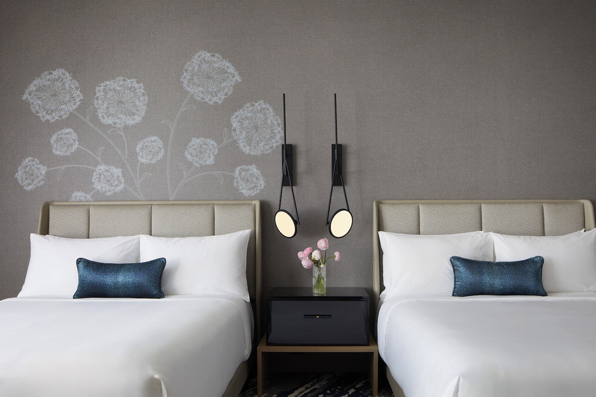 A hotel room with two beds, a nightstand with a vase of flowers, and wall-mounted lamps.