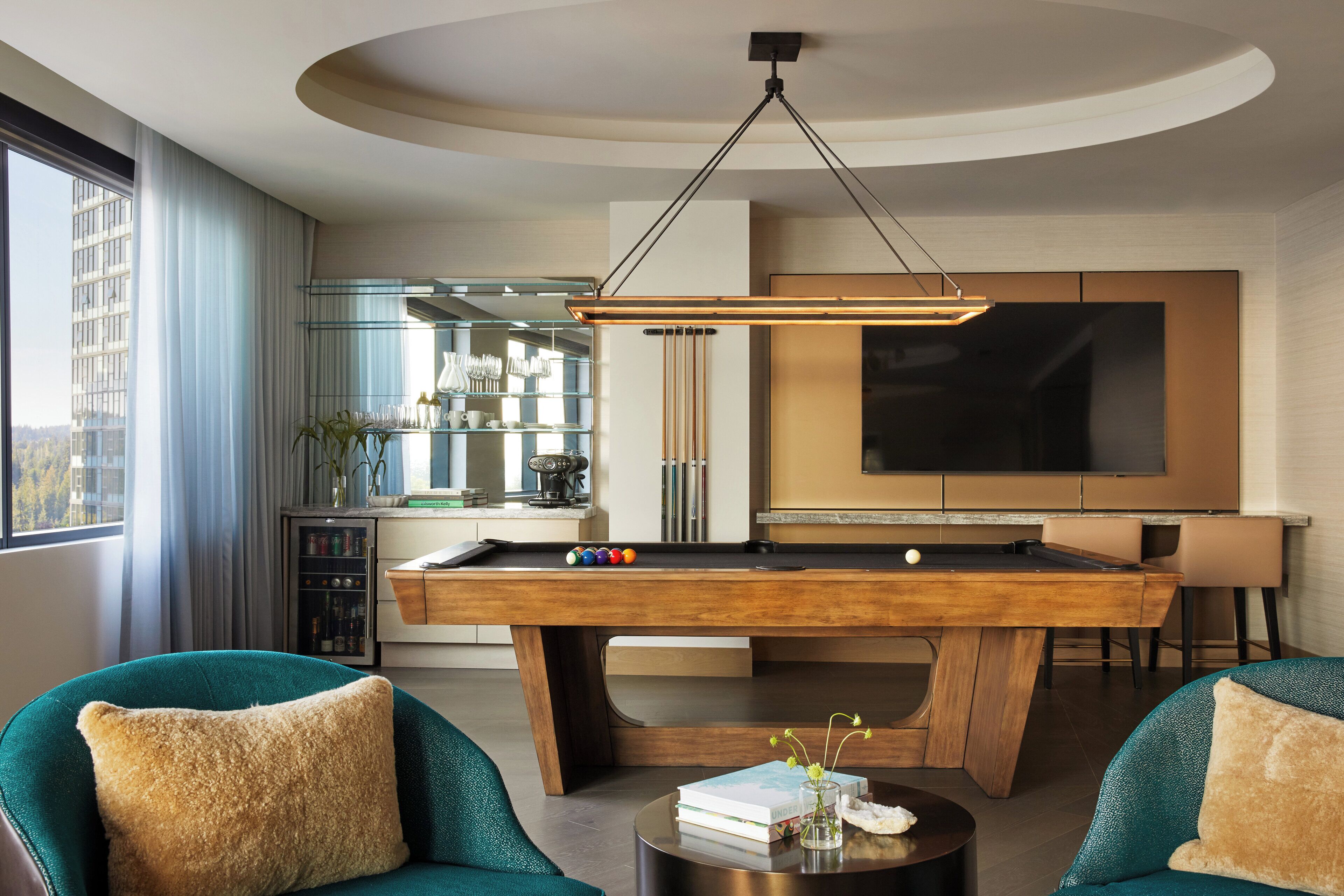 Una sala de estar moderna con mesa de billar, sillones color aguamarina, una mesa de centro con libros y un florero, y un televisor grande en la pared.