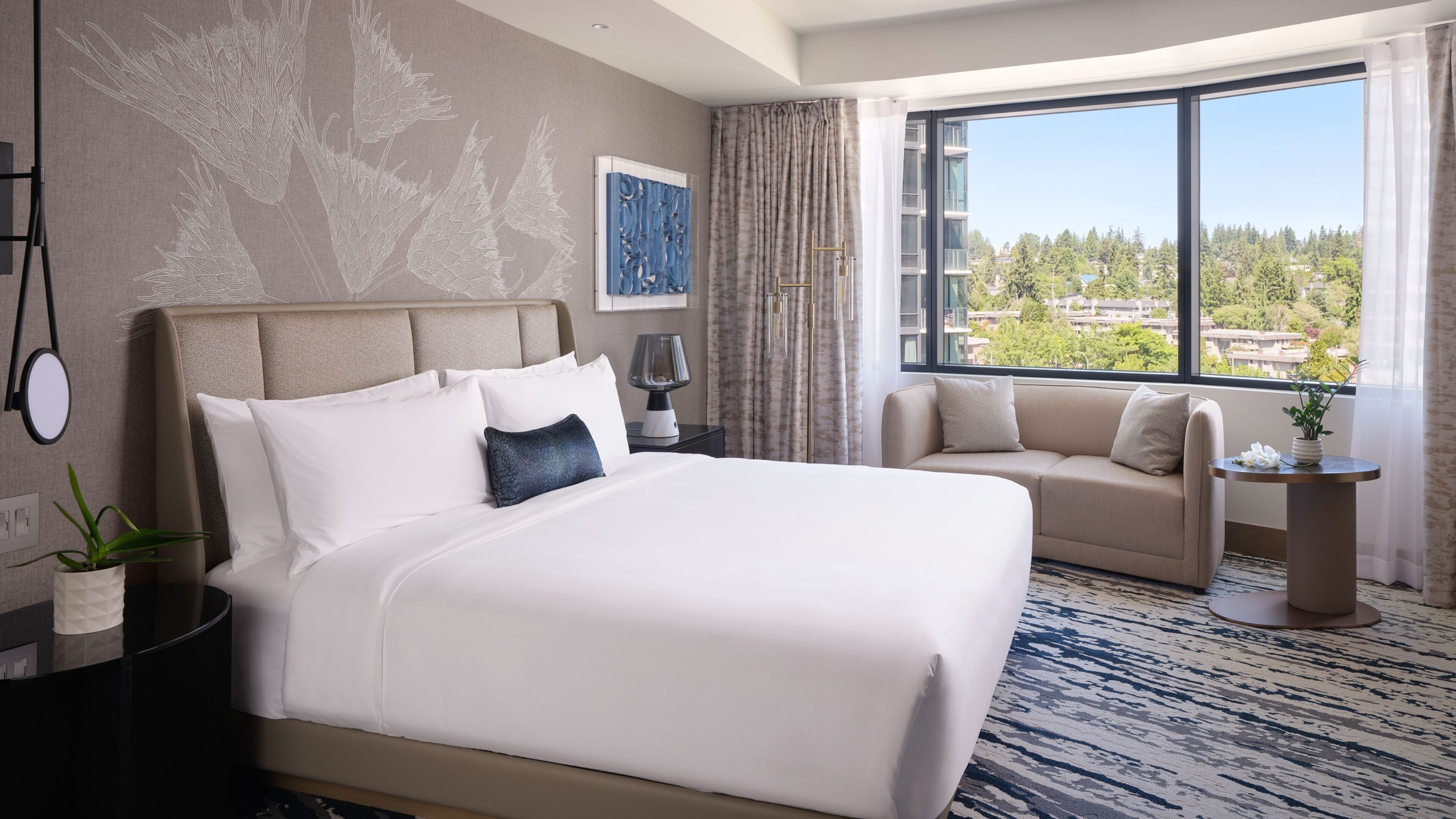 A hotel room with a large bed, a sofa, a nightstand, a lamp, a plant, and a window with a city view.