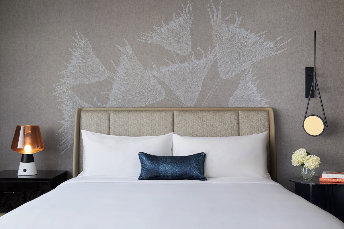 A hotel room with a bed, a nightstand, a lamp, a wall-mounted mirror, and a wall art piece.