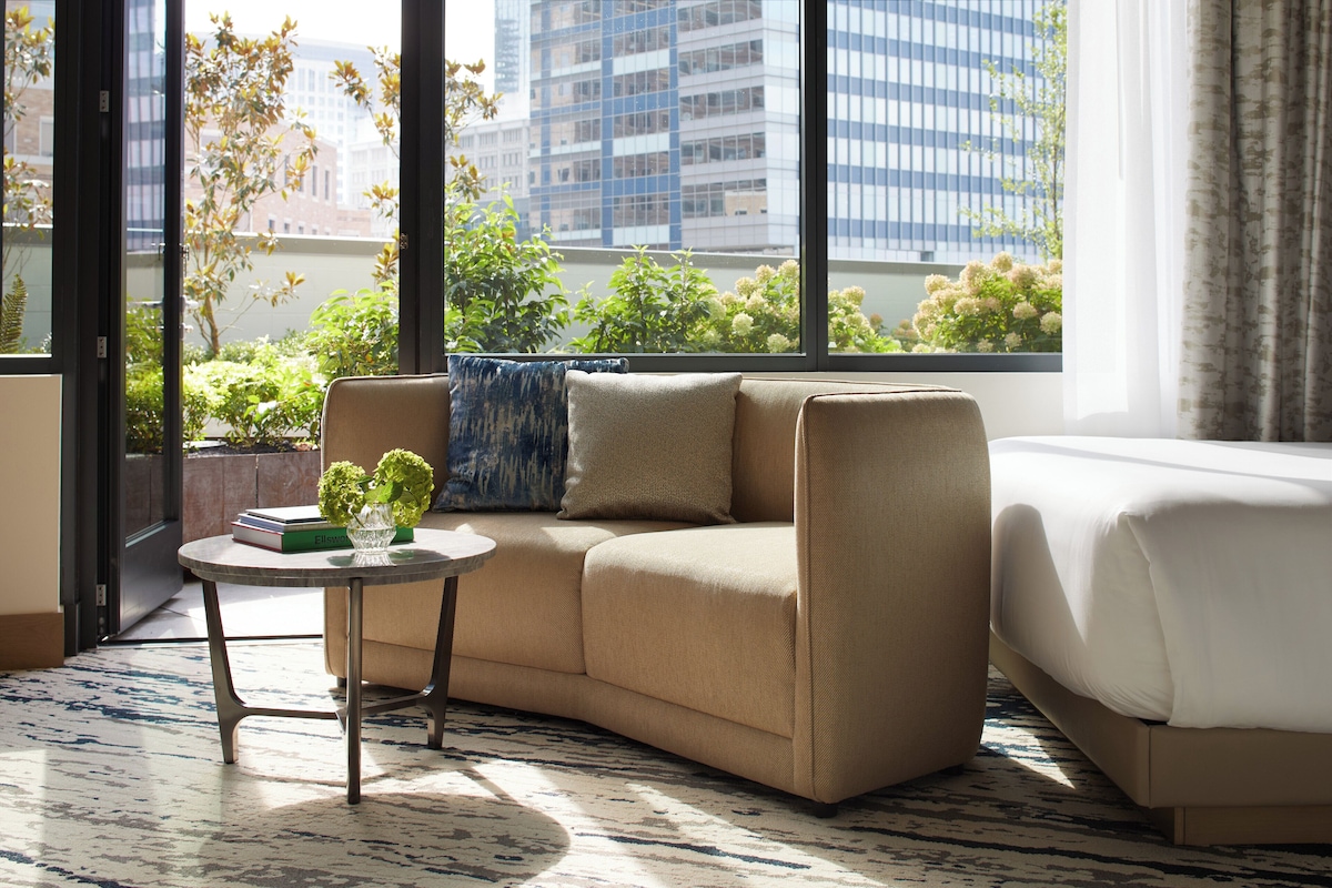 A modern hotel room with a beige sofa, a small round table with a vase of flowers, and a large window offering a view of city buildings and greenery.