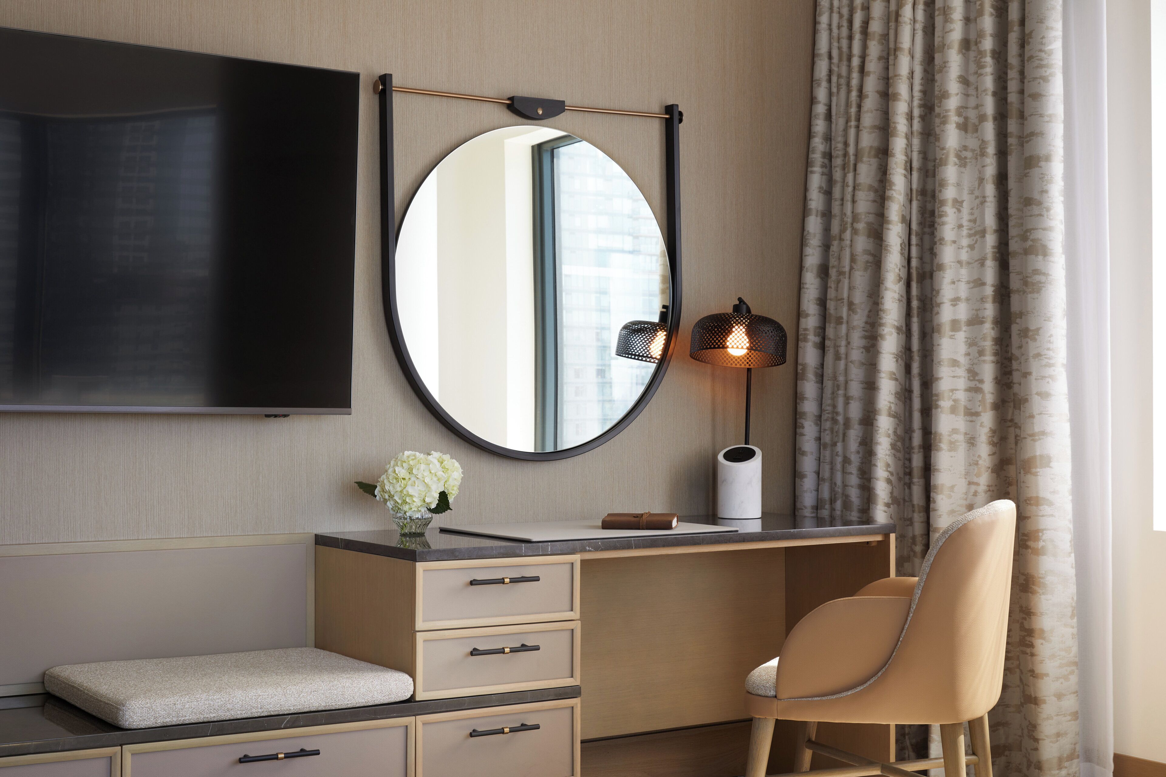Een moderne hotelkamer met een flatscreen televisie aan de muur, een ronde spiegel, een bureau met lades, een gestoffeerde bank, een stoel, een lamp en een raam met gordijnen.