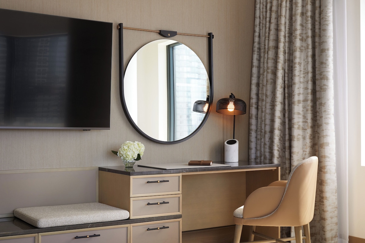 A modern hotel room with a flat-screen TV mounted on the wall, a round mirror, a desk with drawers, a cushioned bench, a chair, a lamp, and a window with curtains.