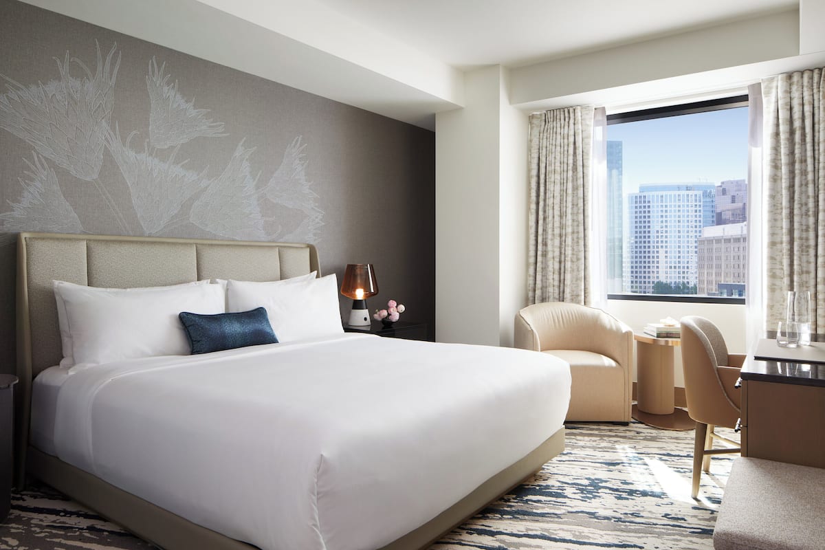 A hotel room with a large bed, a desk, a chair, and a view of the city through the window.