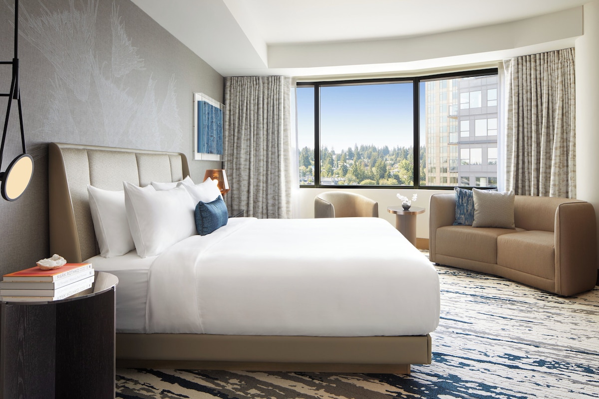 A modern hotel room with a large bed, a sofa, a small table, and a view of a cityscape.