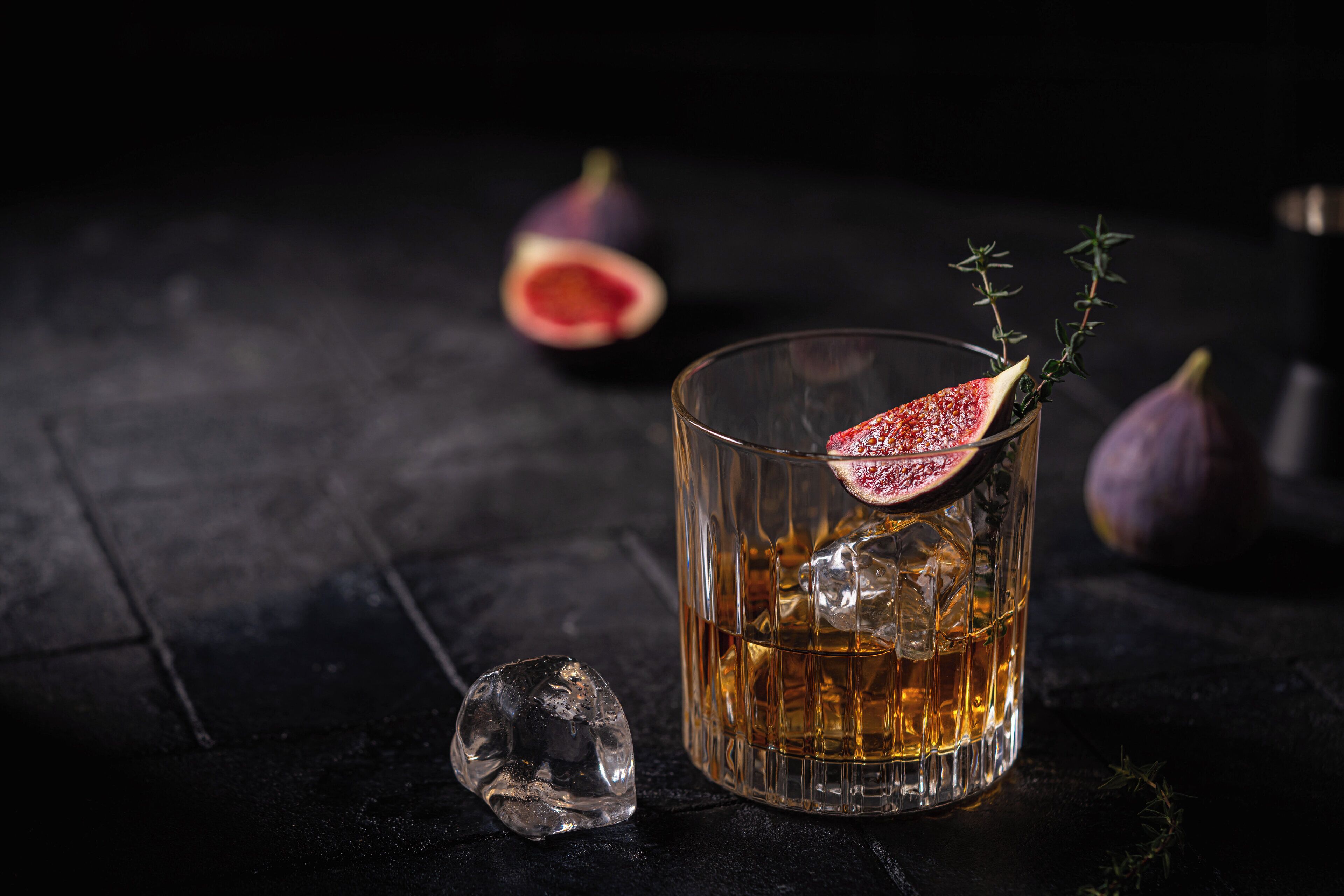 A glass of amber liquid with ice cubes, garnished with a slice of fruit and a sprig of herb, on a dark surface.