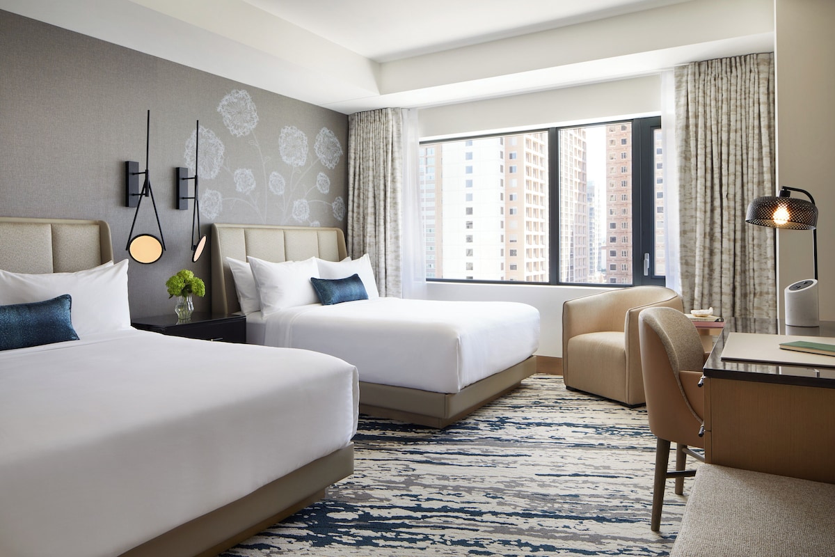 A hotel room with two beds, a desk, a chair, a large window with a city view, and a patterned rug.