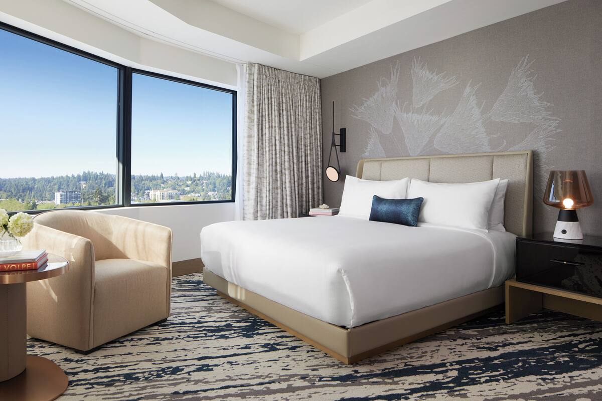 A hotel room with a large bed, a chair, a nightstand, a lamp, and a view of the city.