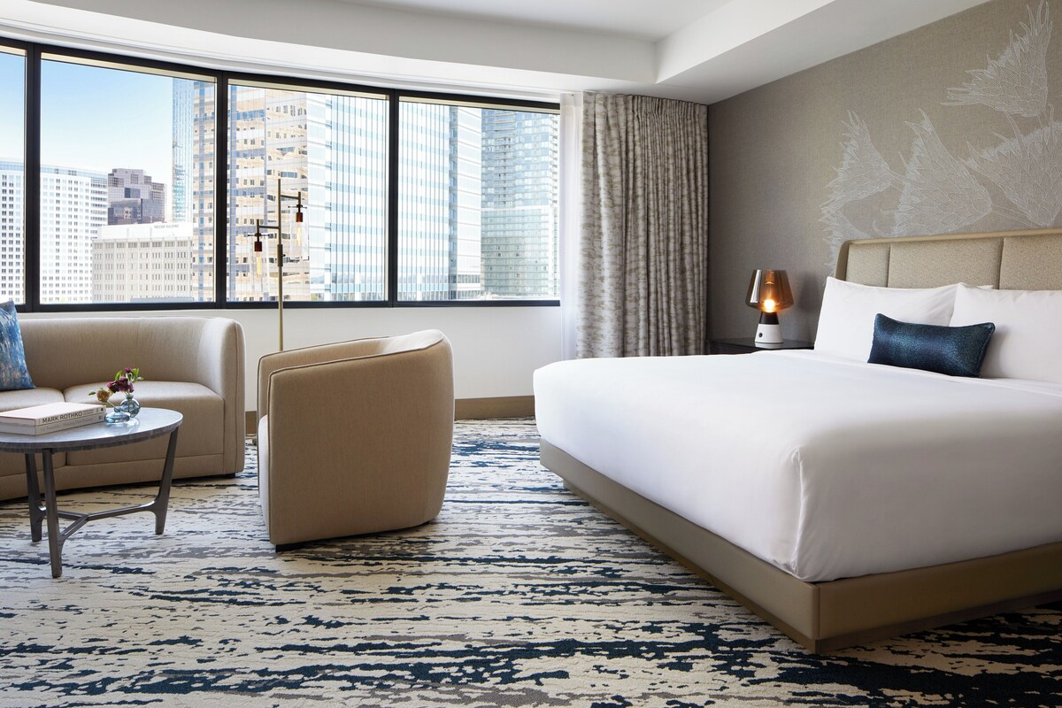A hotel room with a large bed, a sofa, a small table with a lamp, and a view of city buildings through large windows.