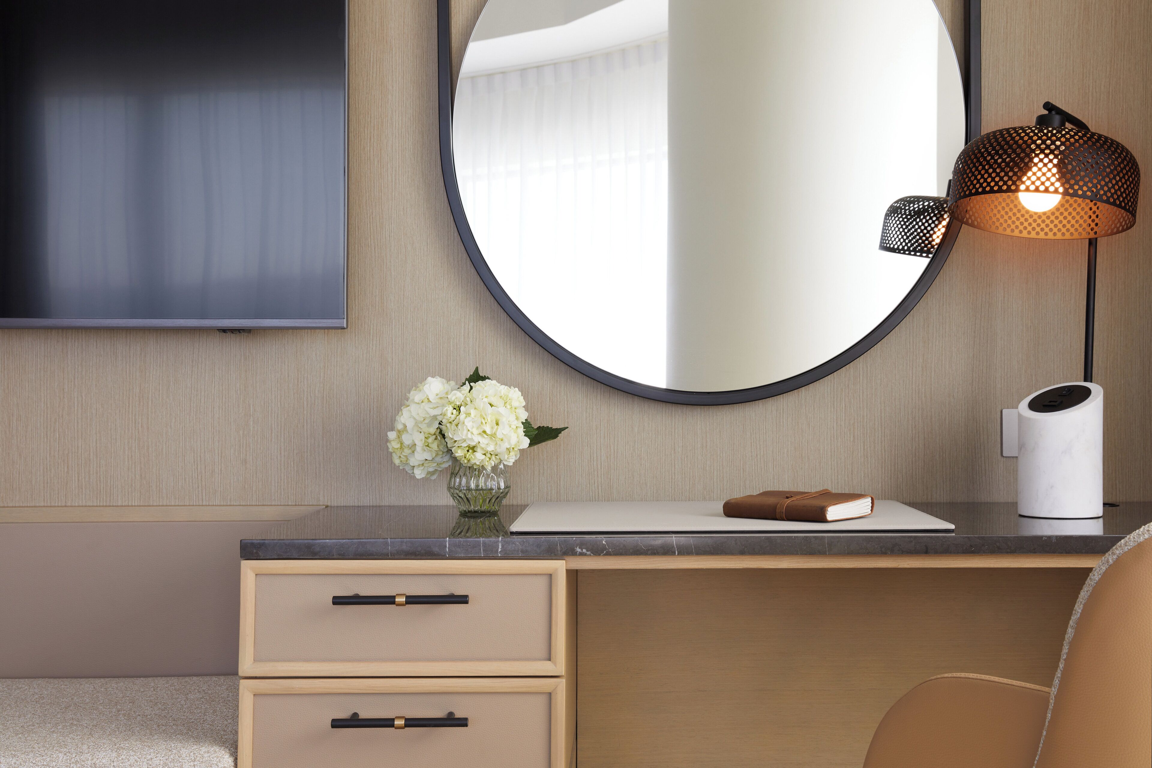 A modern hotel room with a wooden desk, a round mirror, a vase with flowers, a laptop, a book, and a lamp.