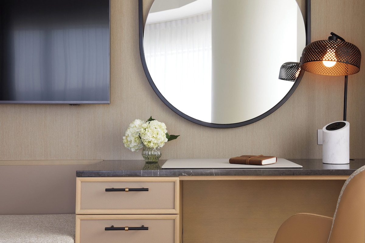 A modern hotel room with a wooden desk, a round mirror, a vase with flowers, a laptop, a book, and a lamp.
