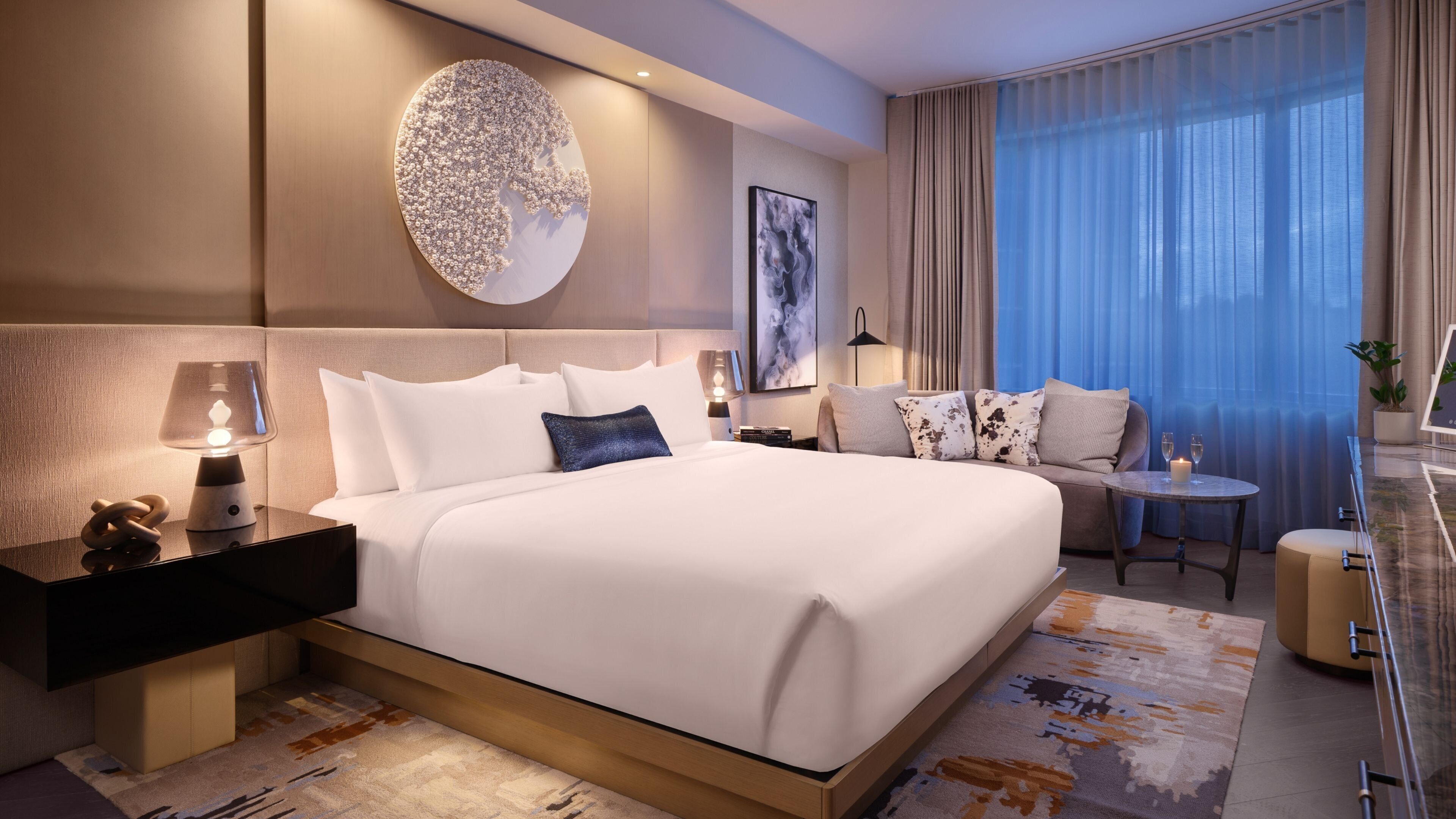 A modern hotel room with a large bed, a bedside table with a lamp, a small round table with a candle, and a view of the city through large windows.