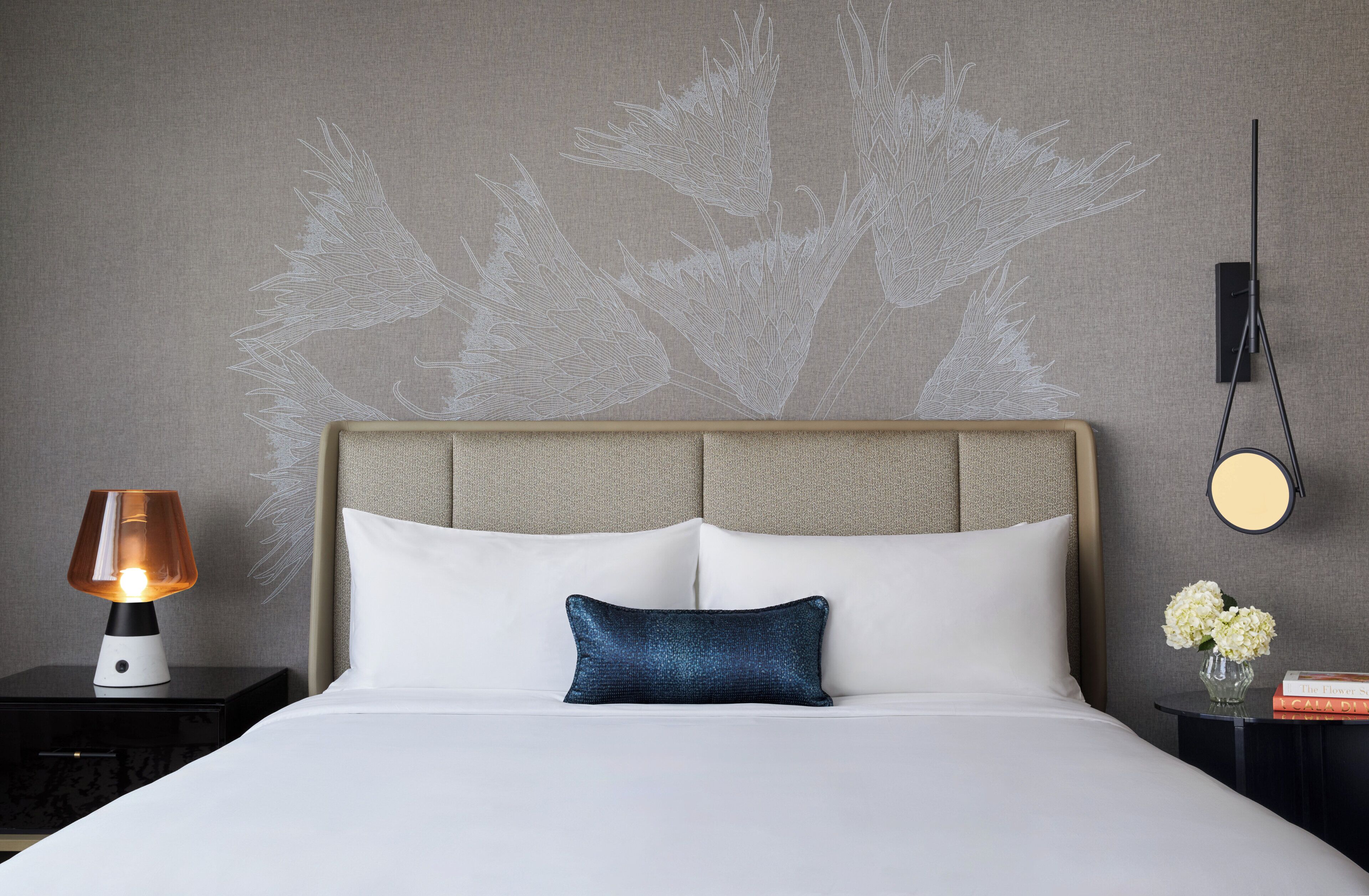 A hotel room with a bed, a nightstand, a lamp, a wall-mounted mirror, and a wall art piece.