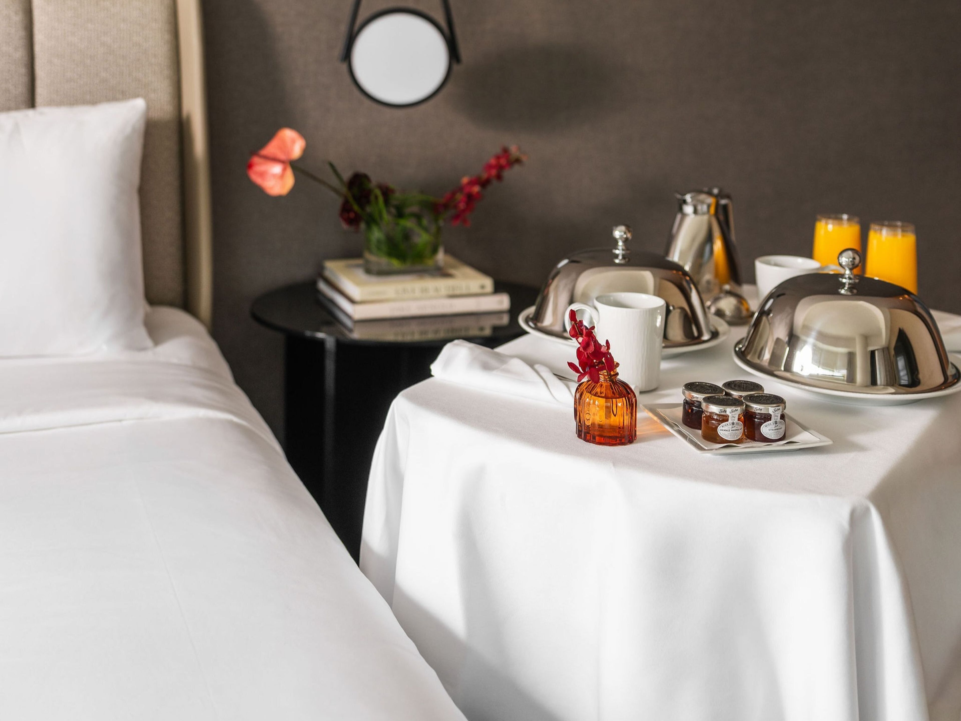 A neatly made bed with a white bedspread, a small table with a white tablecloth, a vase with flowers, a tray with jam jars, a teapot, a cup, and a book.