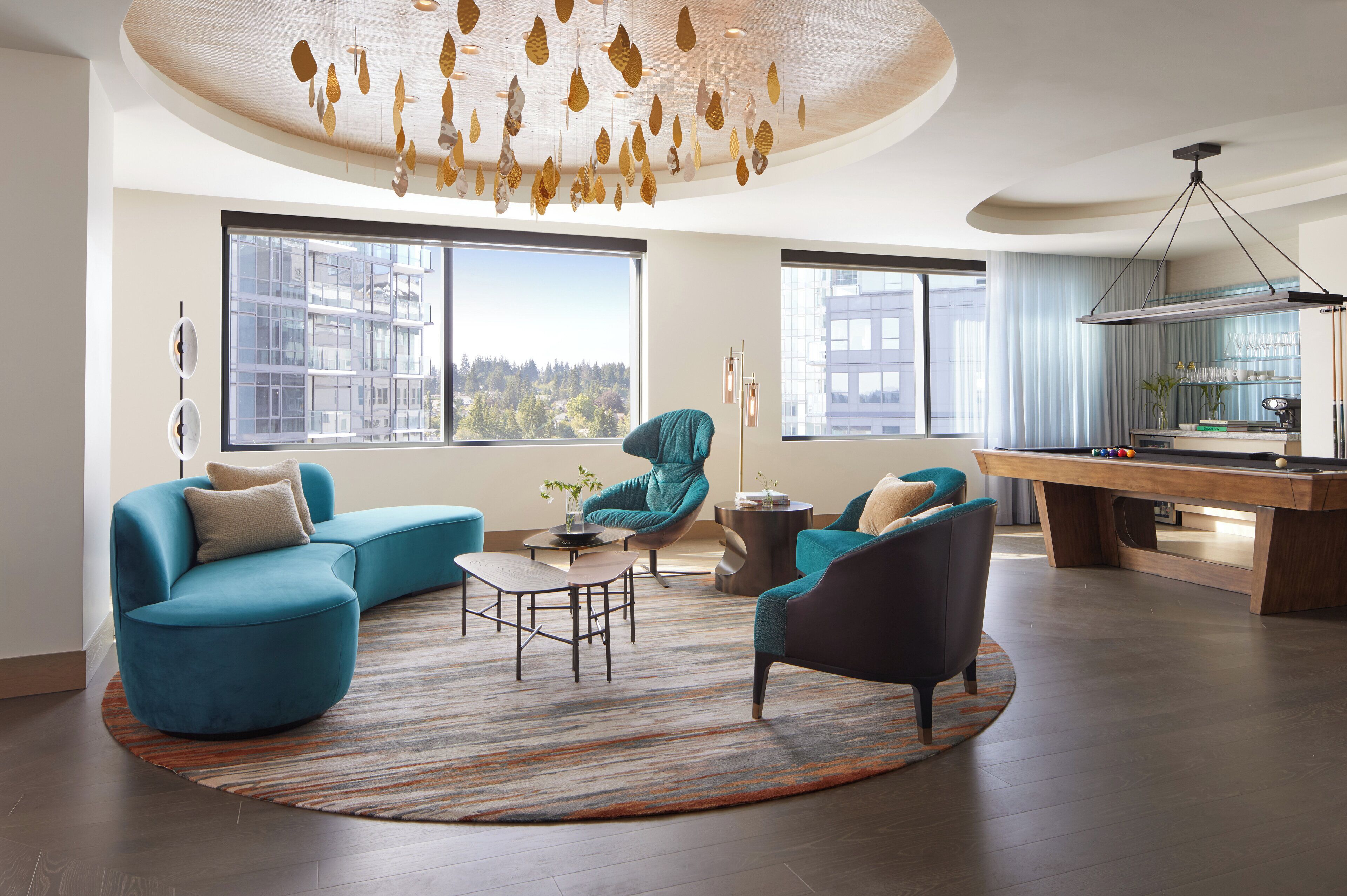 A modern living room with a pool table, teal sofas, and a large window with a city view.