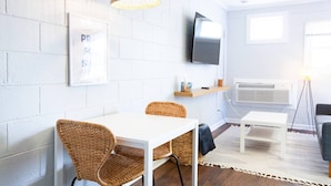 A modern living room with a white brick wall, a flat-screen TV mounted above a wooden shelf, wicker chairs, a white table, and a floor lamp. - Steps To Beach, Kure Pier, Restaurants, & More! (Kure Beach)