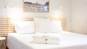 A neatly made bed with white linens and pillows, a framed picture of a surfer on the wall, and a stack of folded towels on the bed. - Steps To Beach, Kure Pier, Restaurants, & More! (Kure Beach)