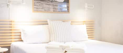 A neatly made bed with white linens and pillows, a framed picture of a surfer on the wall, and a stack of folded towels on the bed.