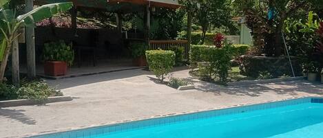 A clear blue pool with a wooden deck and a covered seating area surrounded by lush greenery and trees.