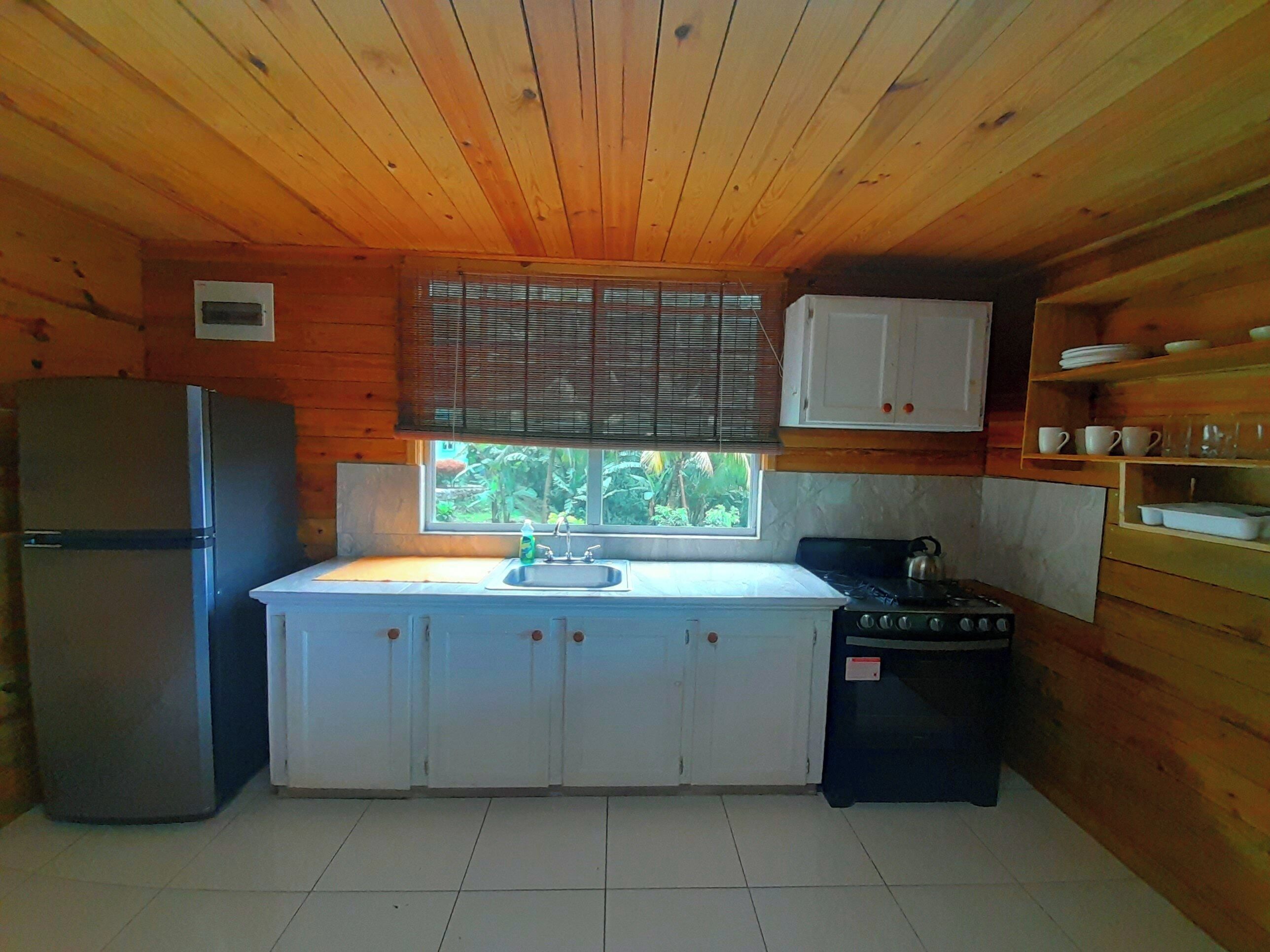 A compact kitchen with wooden walls, a refrigerator, a stove, a sink, and a window with blinds.