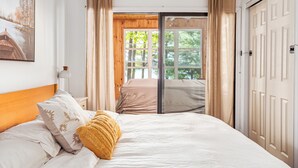 A bedroom with a bed, a nightstand, a lamp, a window with curtains, and a door leading to an outdoor area.