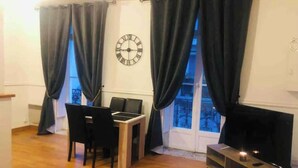 A living room with a wooden floor, a dining table with chairs, a TV on a stand, and a large window with curtains.